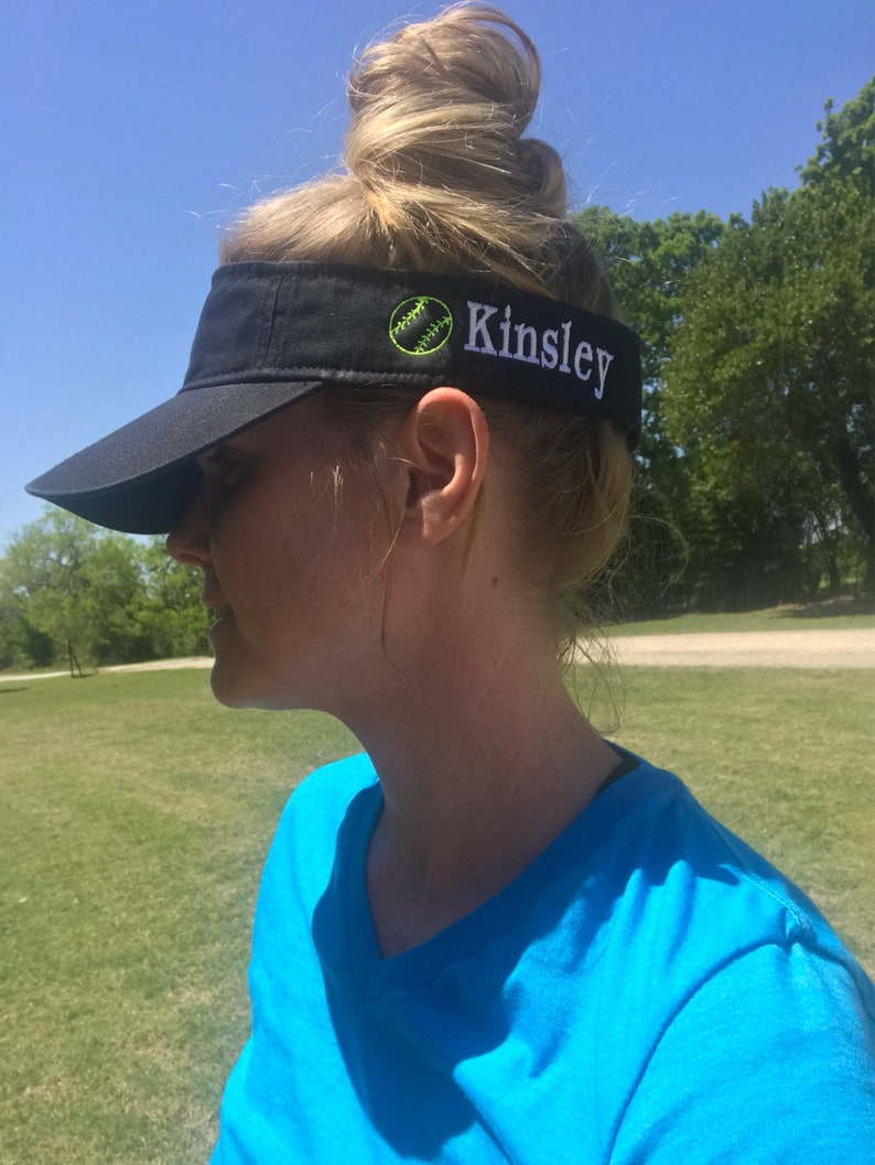 May include: Black visor with a curved brim. The visor has a softball graphic and the name "Kinsley" embroidered in white. The visor is worn by a person wearing a blue v-neck shirt.