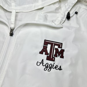 May include: White hooded jacket with a full zipper and the embroidered maroon and black "A&M Aggies" logo on the chest. The jacket has a drawstring hood and a lightweight design.