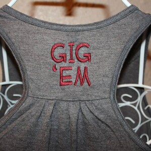 May include: Close-up of a gray tank top with the embroidered words "GIG 'EM" in red. The tank top is hanging on a white wire hanger. The racerback style top is made of a soft material.