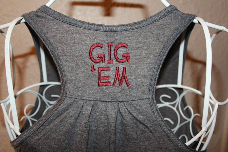 May include: Close-up of a gray tank top with the embroidered words "GIG 'EM" in red. The tank top is hanging on a white wire hanger. The racerback style top is made of a soft material.