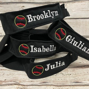May include: Black baseball visors embroidered with names and baseball graphics. The names "Brooklyn", "Giuliana", "Isabella", and "Julia" are displayed in white, along with a baseball design in red and yellow.