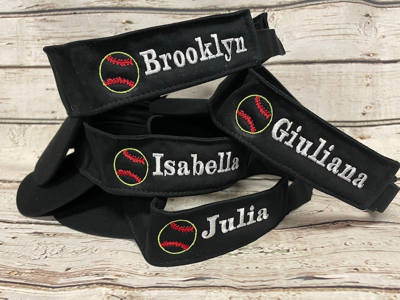 May include: Black baseball visors embroidered with names and baseball graphics. The names "Brooklyn", "Giuliana", "Isabella", and "Julia" are displayed in white, along with a baseball design in red and yellow.