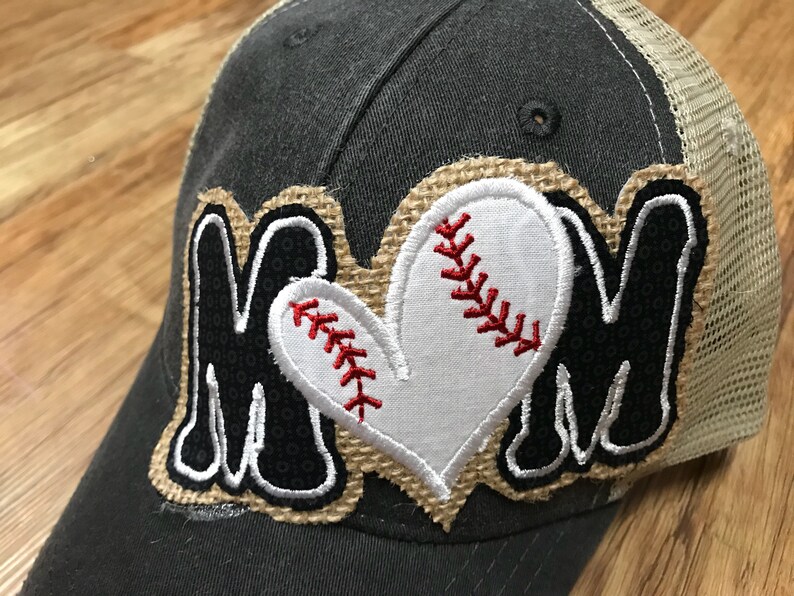 May include: A baseball cap with a dark gray crown and a tan mesh back. The cap features a patch with the word "MOM" in black letters, a white baseball heart, and red stitching.