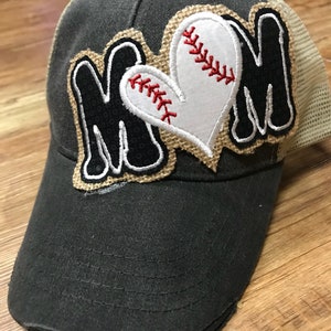 May include: A distressed gray baseball cap with a mesh back. The cap features the word "MOM" in black letters with a baseball heart design. The heart is white with red stitching, and the design is outlined with a burlap-like material.