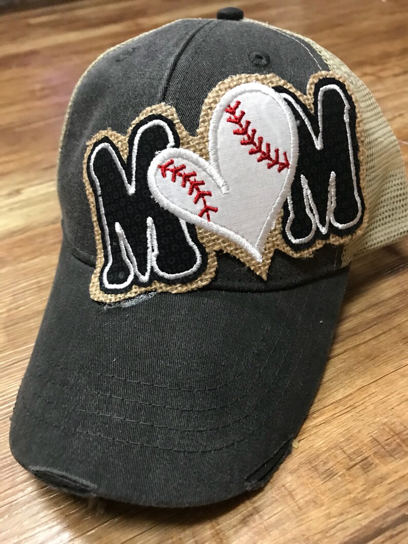 May include: A distressed gray baseball cap with a mesh back. The cap features the word "MOM" in black letters with a baseball heart design. The heart is white with red stitching, and the design is outlined with a burlap-like material.