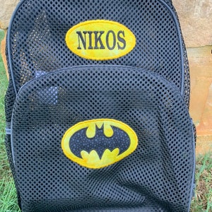 May include: Black mesh backpack with a Batman logo and the name "NIKOS" in yellow. The backpack has a large main compartment and a smaller front pocket. The Batman logo is yellow with a black bat symbol.