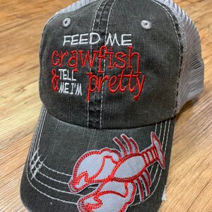 May include: Distressed black baseball cap with a gray mesh back. The front features red embroidered text that reads "FEED ME crawfish & TELL ME I'M pretty." A gray and red crawfish patch is on the brim.