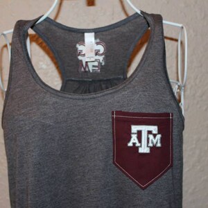 May include: Gray tank top with a maroon pocket featuring the letters "A" "T" and "M" in white. The tank top has a racerback design and a tag with text on the back.