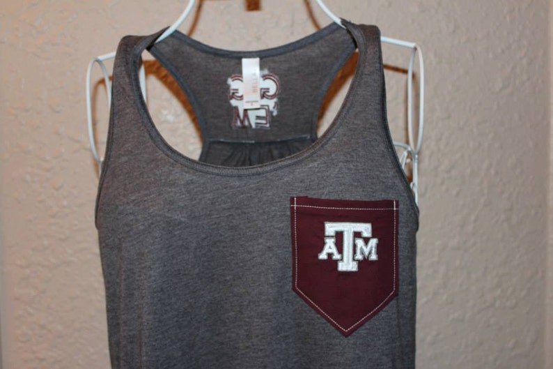 May include: Gray tank top with a maroon pocket featuring the letters "A" "T" and "M" in white. The tank top has a racerback design and a tag with text on the back.