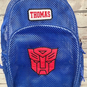 May include: Blue mesh backpack with the name "THOMAS" embroidered on a patch. The backpack features a red Transformers logo and a zippered closure. The bag has adjustable straps and is designed for carrying items.