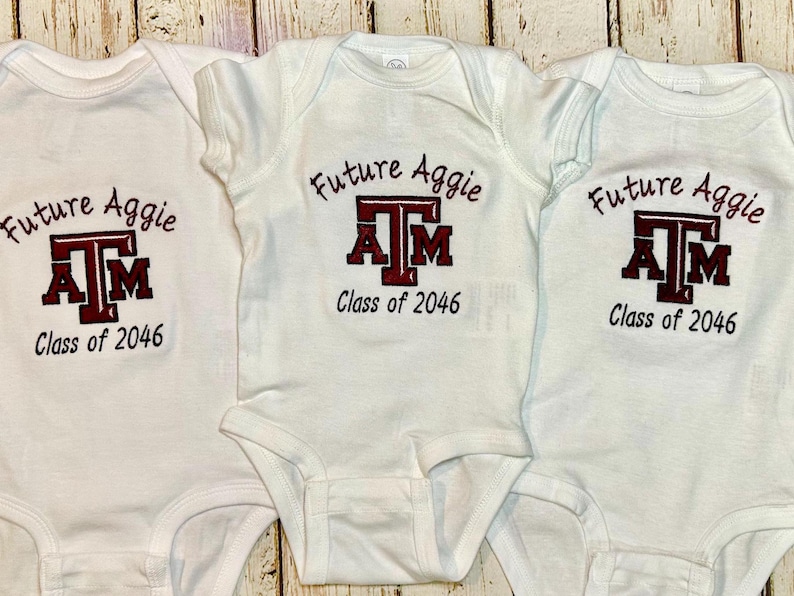 May include: Three white baby onesies with the text "Future Aggie" above a maroon and white "ATM" logo and the words "Class of 2046" below. The onesies are made of soft material.