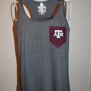 May include: A gray tank top with a maroon pocket featuring the letters "A" and "M" with a "T" in the middle. The tank top has a racerback design and a scoop neck. The brand name is printed on the back of the tank top.