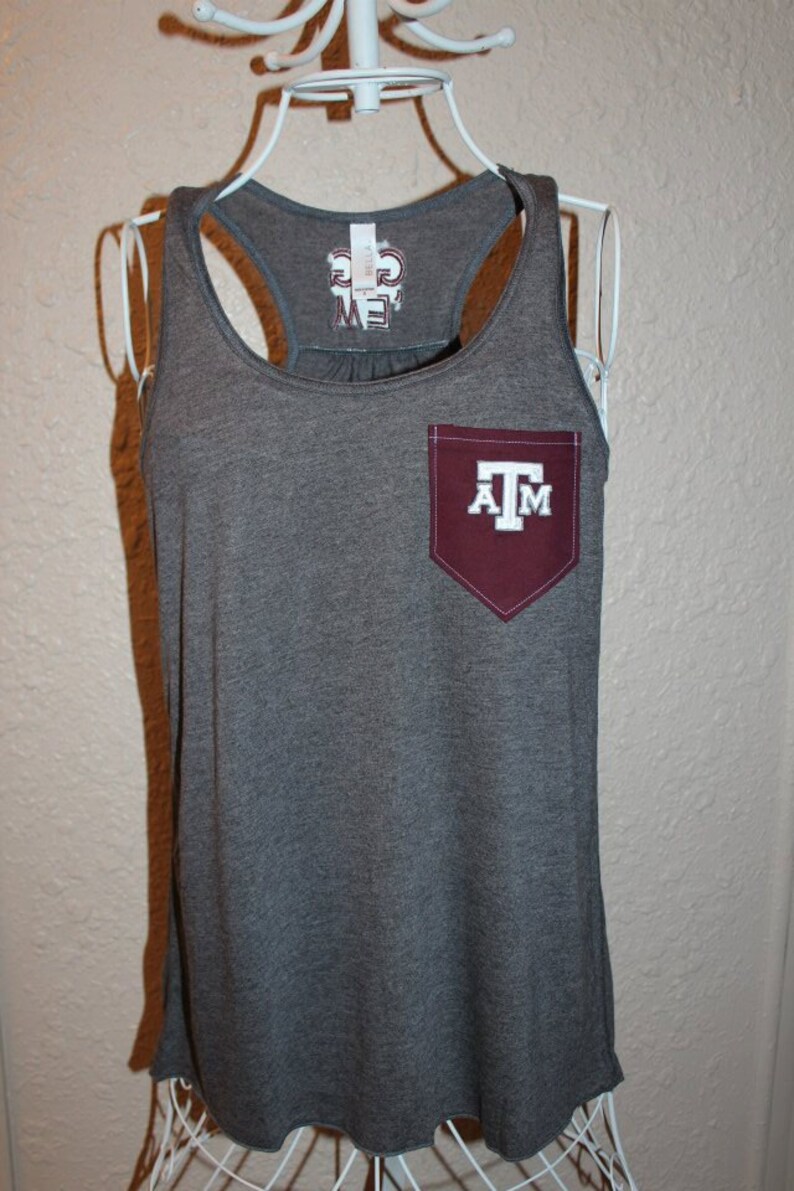 May include: A gray tank top with a maroon pocket featuring the letters "A" and "M" with a "T" in the middle. The tank top has a racerback design and a scoop neck. The brand name is printed on the back of the tank top.