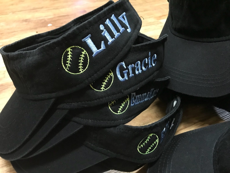 May include: Black visors embroidered with names in light blue thread and a yellow softball design. The names "Lilly", "Gracie", and "Emma" are visible. Several visors are stacked together.