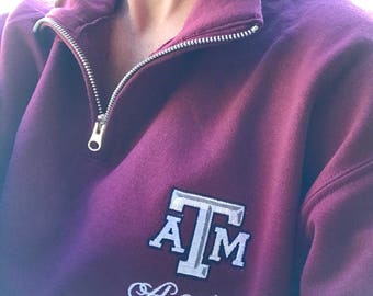 May include: A maroon quarter-zip sweatshirt with a silver zipper. The sweatshirt features the letters "A T M" in a white and gray design, with the word "Aggies" embroidered below in a cursive font. The collar is folded down.