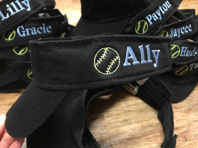 May include: Black visor hats embroidered with names and a softball design. The name "Ally" is embroidered in light blue, with a yellow softball graphic. Other names like "Lilly", "Gracie", "Payton", "Jaycee", "Hudson", and "Taylor" are also visible.