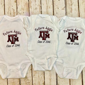 May include: Three white baby onesies with the text "Future Aggie" and "Class of 2046" printed in maroon and black. The Texas A&M logo is also featured on the front of each onesie.