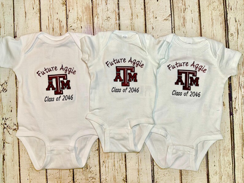 May include: Three white baby onesies with the text "Future Aggie" and "Class of 2046" printed in maroon and black. The Texas A&M logo is also featured on the front of each onesie.