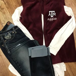May include: A maroon fleece vest with the text "ATM aggies" is displayed over a white long-sleeved shirt, paired with dark wash jeans. The jeans have decorative stitching and a light blue cuff. The outfit is arranged on a wooden surface.