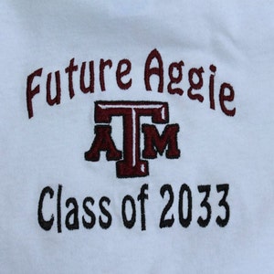 May include: White onesie with embroidered text. The text reads "Future Aggie" in maroon above a maroon and white "ATM" logo, with "Class of 2033" in black below. The onesie is made of soft fabric.