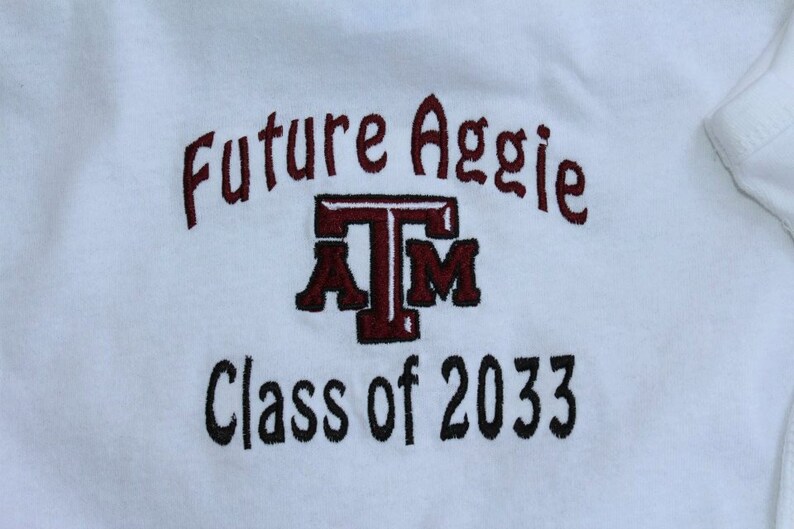 May include: White onesie with embroidered text. The text reads "Future Aggie" in maroon above a maroon and white "ATM" logo, with "Class of 2033" in black below. The onesie is made of soft fabric.