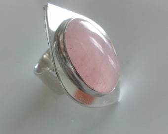 May include: A silver ring with a large oval-shaped pink gemstone set in a silver bezel. The ring band is wide and has a unique, asymmetrical design.