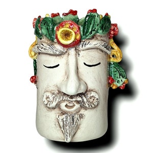 Könnte beinhalten: Eine cremefarbene Keramik-Gesichtsskulptur mit geschlossenen Augen, Schnurrbart und Bart. Die Skulptur hat grüne, rote und gelbe Akzente. Eine Blätterkrone ziert den oberen Teil des Kopfes.
