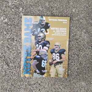 May include: A printed program for a Notre Dame vs. Air Force football game. The cover features images of football players in uniform, with the words "Notre Dame" in large blue letters. The program is on a gray concrete surface.