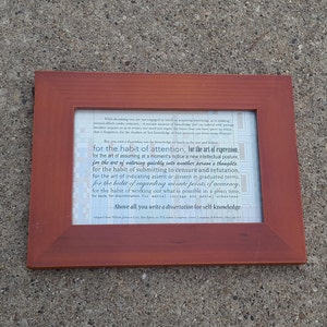 May include: A rectangular, brown wooden picture frame with a white printed document inside. The document features black text in a serif font, with the phrase "for the habit of attention, for the art of expression" highlighted. The frame is resting on a gray surface.