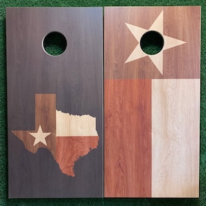 K&ouml;nnte beinhalten: Zwei h&ouml;lzerne Cornhole-Bretter mit einem Texas-Flaggen-Design. Ein Brett zeigt eine Silhouette des Bundesstaates Texas mit einem Stern in der Mitte, w&auml;hrend das andere Brett eine traditionelle Texas-Flagge mit einem wei&szlig;en Stern auf blauem Hintergrund zeigt.