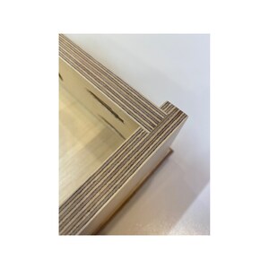 May include: Close-up of a corner of a wooden box made of plywood. The wood is light brown and has a natural grain pattern. The box is made of multiple layers of wood, which are visible in the corner.
