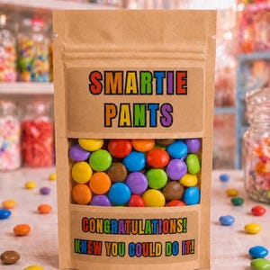 Könnte beinhalten: Ein brauner Papierbeutel gefüllt mit bunten, schokolierten Bonbons. Das Etikett zeigt den Text SMARTIE PANTS in Regenbogenfarben, mit CONGRATULATIONS! KNEW YOU COULD DO IT! in einem passenden Stil unterhalb des Sichtfensters gedruckt.