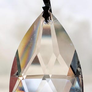 Puede incluir: Un colgante de cristal transparente facetado en forma de l&aacute;grima, suspendido por una peque&ntilde;a cuenta negra y una pieza de cristal octogonal en la parte superior. Las superficies de vidrio presentan cortes geom&eacute;tricos que refractan la luz.