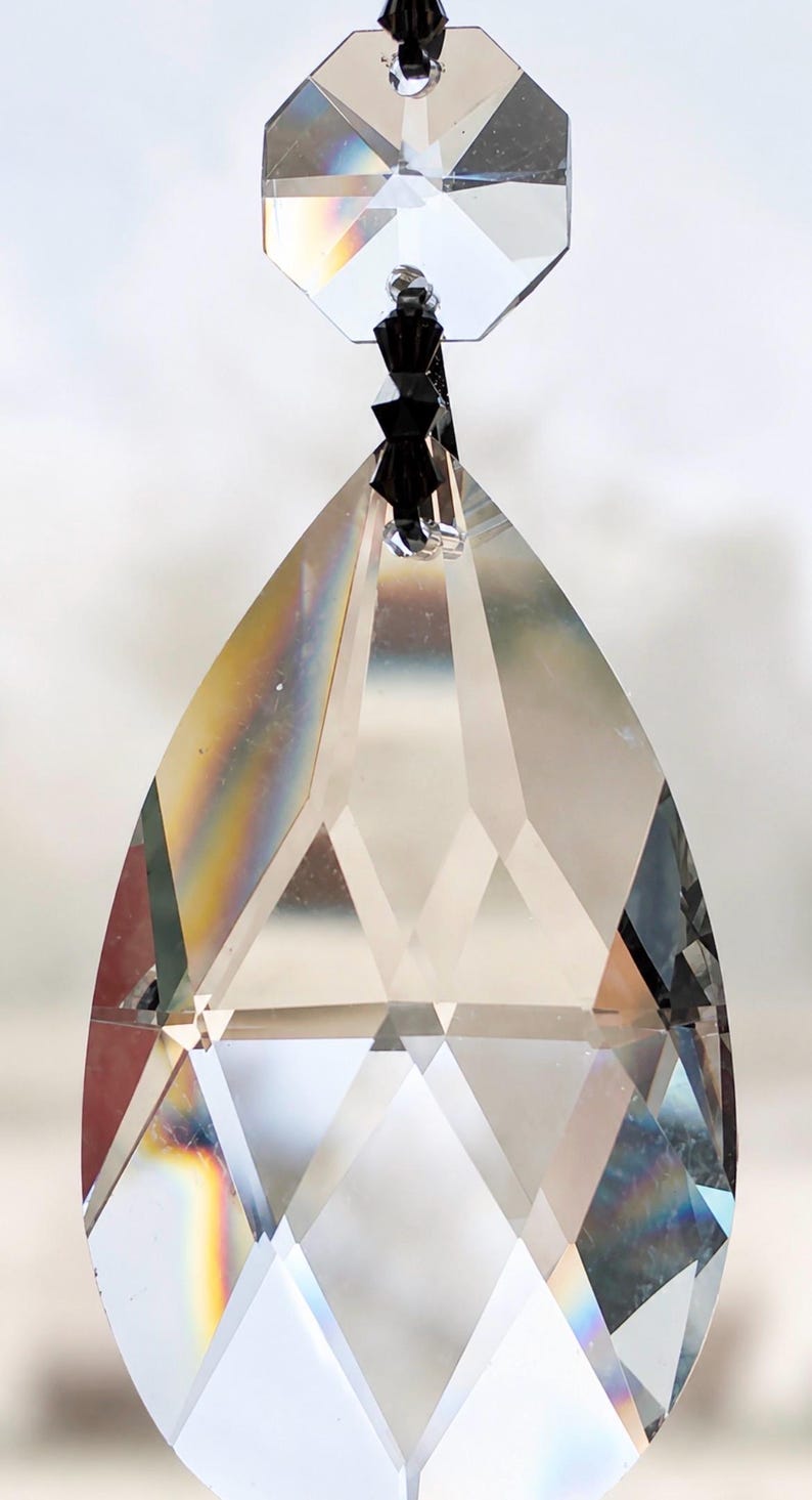 Puede incluir: Un colgante de cristal transparente facetado en forma de l&aacute;grima, suspendido por una peque&ntilde;a cuenta negra y una pieza de cristal octogonal en la parte superior. Las superficies de vidrio presentan cortes geom&eacute;tricos que refractan la luz.
