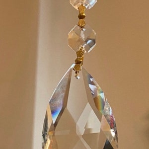 May include: A crystal teardrop pendant with faceted edges hangs from a gold chain with smaller crystal beads. The pendant is clear and reflects light.