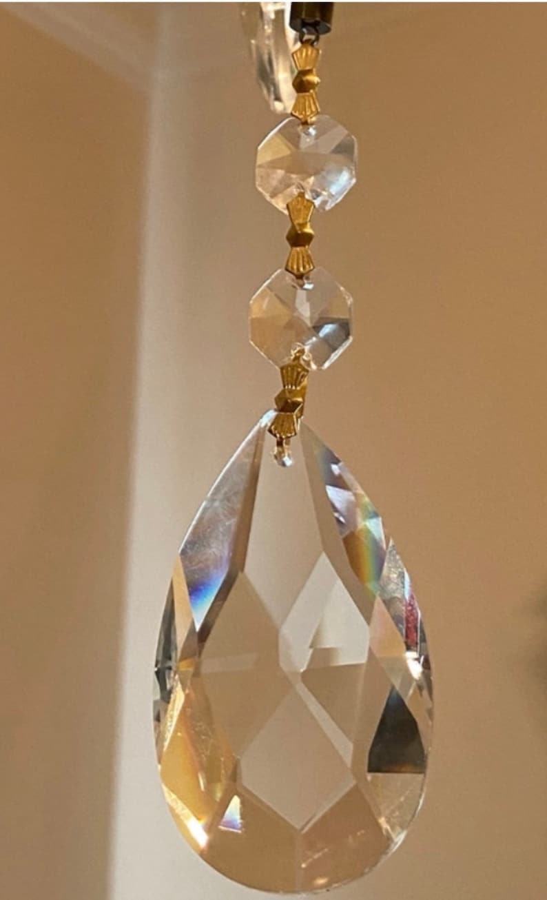 May include: A crystal teardrop pendant with faceted edges hangs from a gold chain with smaller crystal beads. The pendant is clear and reflects light.