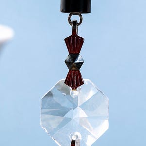 Puede incluir: Un adorno colgante decorativo con un colgante de cristal octogonal facetado y transparente, suspendido por un anillo de metal plateado. Sobre el cristal hay dos cuentas de color rojo oscuro en forma de abanico separadas por una cuenta met&aacute;lica.
