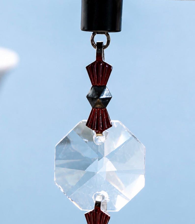 Puede incluir: Un adorno colgante decorativo con un colgante de cristal octogonal facetado y transparente, suspendido por un anillo de metal plateado. Sobre el cristal hay dos cuentas de color rojo oscuro en forma de abanico separadas por una cuenta met&aacute;lica.