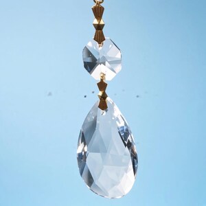 Puede incluir: Un colgante de cristal transparente facetado con una gran gota en forma de l&aacute;grima suspendida bajo un cristal octogonal m&aacute;s peque&ntilde;o. Las dos piezas de vidrio est&aacute;n conectadas por peque&ntilde;os pasadores de lat&oacute;n dorado, creando un adorno colgante.