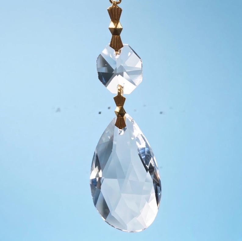 Puede incluir: Un colgante de cristal transparente facetado con una gran gota en forma de l&aacute;grima suspendida bajo un cristal octogonal m&aacute;s peque&ntilde;o. Las dos piezas de vidrio est&aacute;n conectadas por peque&ntilde;os pasadores de lat&oacute;n dorado, creando un adorno colgante.
