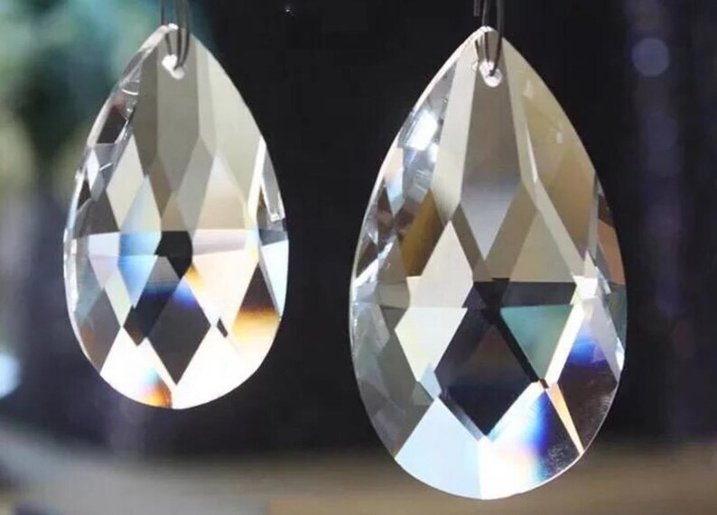 Puede incluir: Dos colgantes de cristal transparente facetado con forma de l&aacute;grima. Cada colgante presenta un peque&ntilde;o orificio en la parte superior para colgarlo y muestra superficies geom&eacute;tricas multifacetadas que refractan la luz con bordes definidos.
