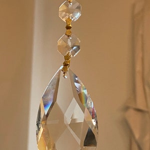 May include: A clear crystal teardrop pendant with gold accents hangs from a chain. The pendant is faceted and reflects light.