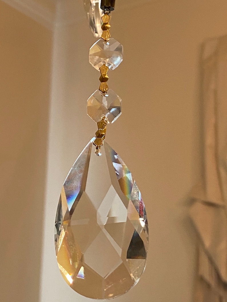 May include: A clear crystal teardrop pendant with gold accents hangs from a chain. The pendant is faceted and reflects light.
