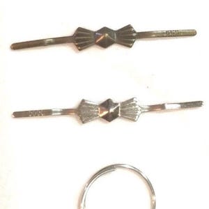 Puede incluir: Tres horquillas de metal dispuestas horizontalmente, cada una con un dise&ntilde;o central en forma de lazo con un centro geom&eacute;trico facetado. Debajo de las horquillas hay un anillo de metal circular. Acabados en tonos dorado, bronce y plateado.