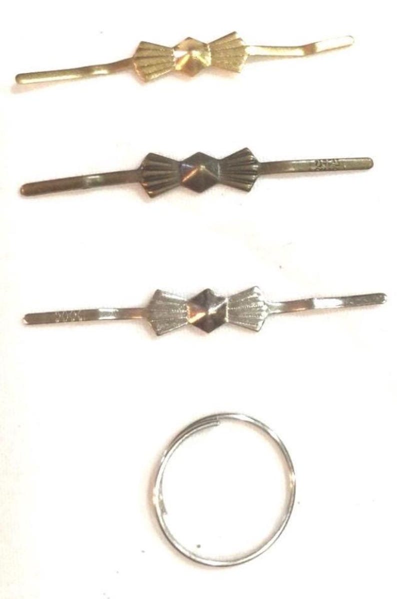 Puede incluir: Tres horquillas de metal dispuestas horizontalmente, cada una con un dise&ntilde;o central en forma de lazo con un centro geom&eacute;trico facetado. Debajo de las horquillas hay un anillo de metal circular. Acabados en tonos dorado, bronce y plateado.