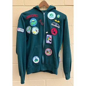 May include: A dark green zip-up hoodie with multiple patches. Patches include "Girl Scouts USA", "Foothills Council", and "Camp Glengarra". The hoodie has a front pocket and a hood. The American Red Cross patch is also visible.