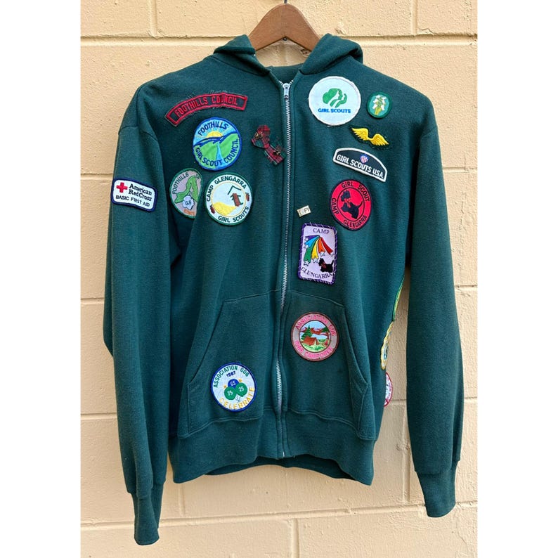 May include: A dark green zip-up hoodie with multiple patches. Patches include "Girl Scouts USA", "Foothills Council", and "Camp Glengarra". The hoodie has a front pocket and a hood. The American Red Cross patch is also visible.