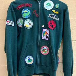 May include: A dark green zip-up hoodie with multiple patches. Patches include "Foothills Council", "Girl Scouts", "Camp Glengarra", and "American Red Cross Basic First Aid". The hoodie has a hood and two front pockets.