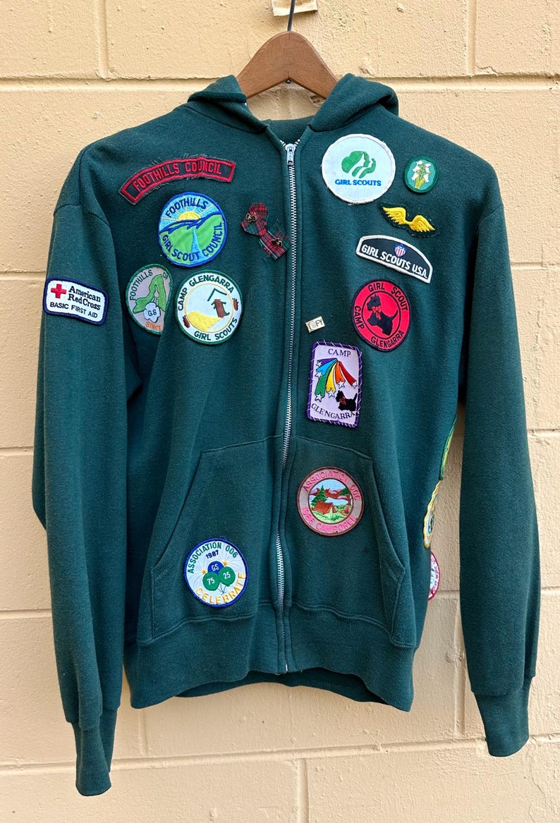 May include: A dark green zip-up hoodie with multiple patches. Patches include "Foothills Council", "Girl Scouts", "Camp Glengarra", and "American Red Cross Basic First Aid". The hoodie has a hood and two front pockets.
