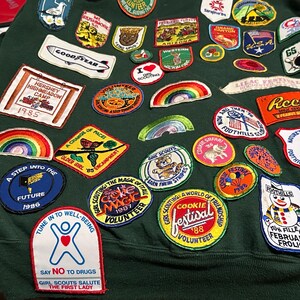 May include: A dark green sweatshirt covered in embroidered patches. Patches include rainbows, a Goodyear logo, and text that reads "Hershey Highmeadow Camp 1985". Other patches say "Cookie Festival" and "Say No To Drugs".